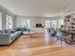 A modern living room with a sectional sofa, dining table, and chairs, a bookshelf, and a TV on a white cabinet.