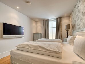 A hotel room with two beds, a TV mounted on the wall, a bedside table with a lamp, and a window with curtains.