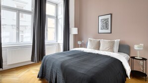 Ein Schlafzimmer mit einem großen Bett, einem Fenster mit Vorhängen, einer Nachttischlampe und einem gerahmten Bild an der Wand.