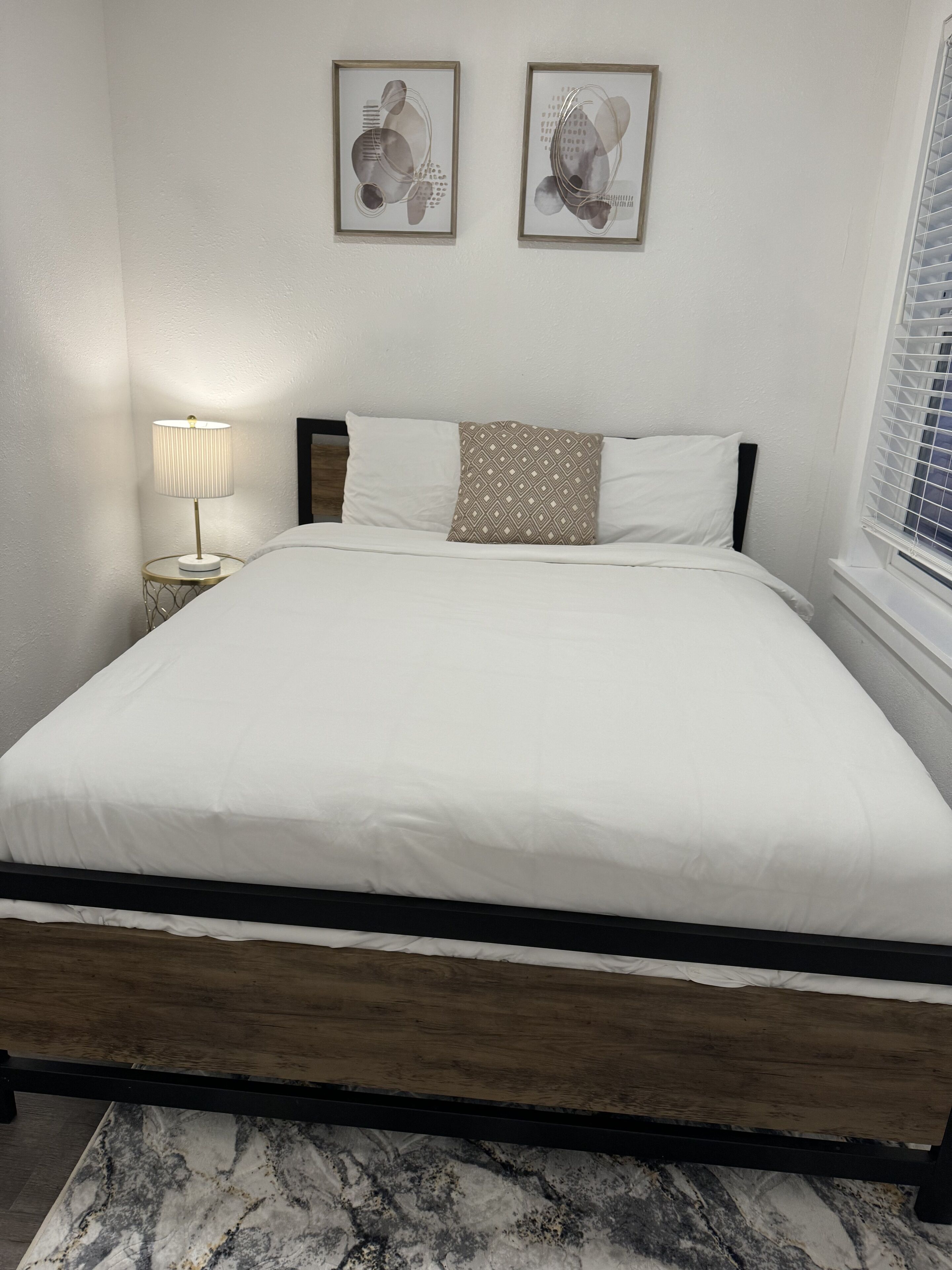 Un lit bien fait, avec une literie blanche, un coussin à motifs et une lampe de chevet. Deux œuvres d’art abstraites, encadrées, sont accrochées au mur au-dessus du lit.