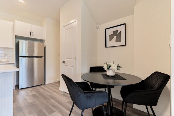 A modern kitchen with white cabinets and a stainless steel refrigerator, a dining area with a round table and black chairs, and a framed abstract art piece on the wall.