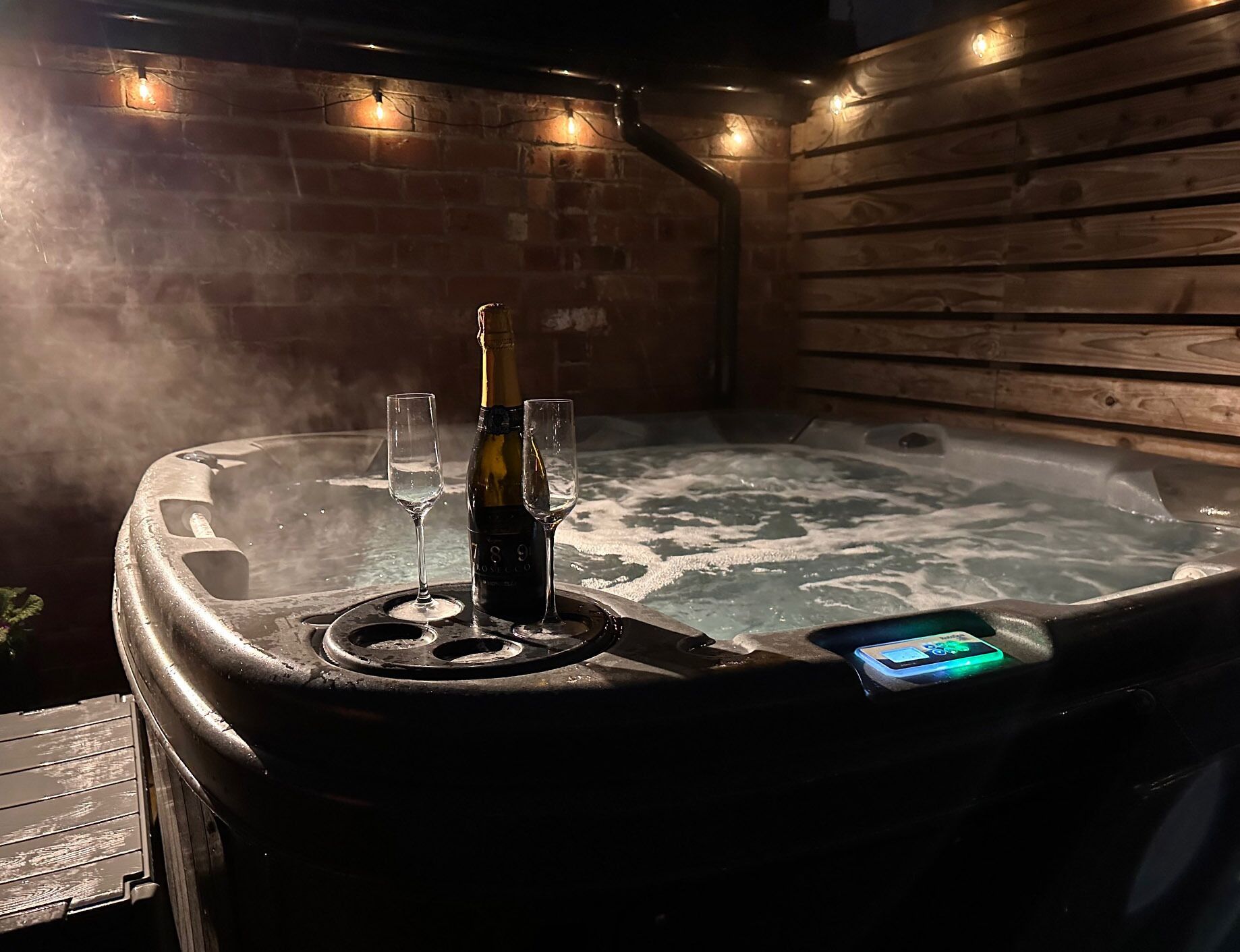A hot tub with two glasses and a bottle of champagne, surrounded by wooden walls and string lights.