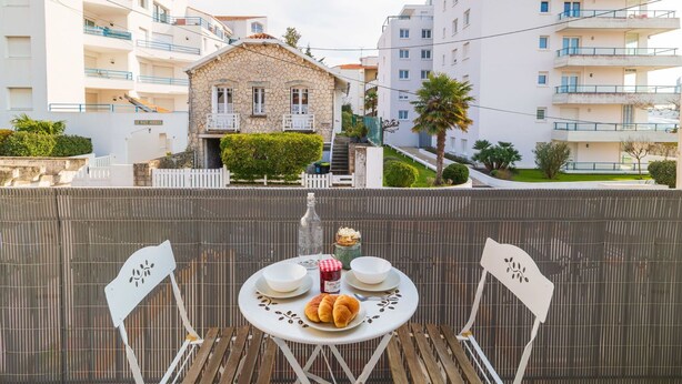 Un balcó amb taula per a dos, amb vista a una zona residencial amb edificis moderns i una casa de pedra.