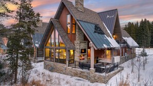 A modern log cabin with large windows, a stone chimney, and a metal roof.