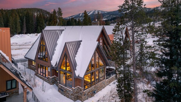 A modern lodge with large windows and a stone facade, surrounded by snow-covered landscape and pine trees.