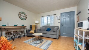 A living room with a sofa, a dining table with chairs, a coffee table, a clock, a door, and a kitchen area with a microwave.