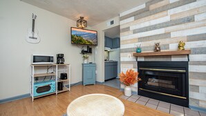 A living room with a fireplace, a television, a guitar, and a kitchen in the background.