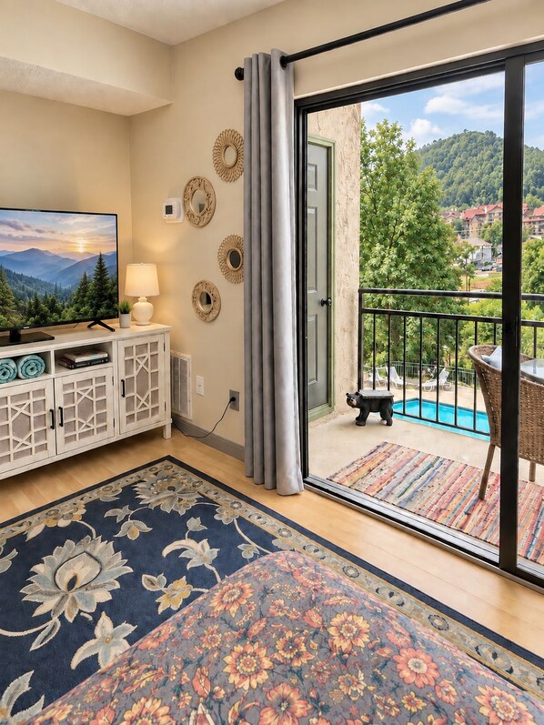 Ein Wohnzimmer mit Fernseher, einem weißen Schrank, einem gemusterten Teppich und einer Schiebetür, die auf einen Balkon mit Blick auf einen Pool führt.