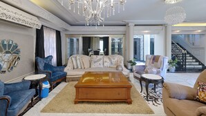 Een ruime woonkamer met een grote houten salontafel, blauwe en beige banken, een kroonluchter en een trap op de achtergrond.