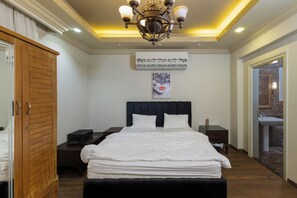 A neatly made bed with white linens, a dark headboard, and a chandelier above. There is a wooden wardrobe to the left and a small table with a lamp to the right.
