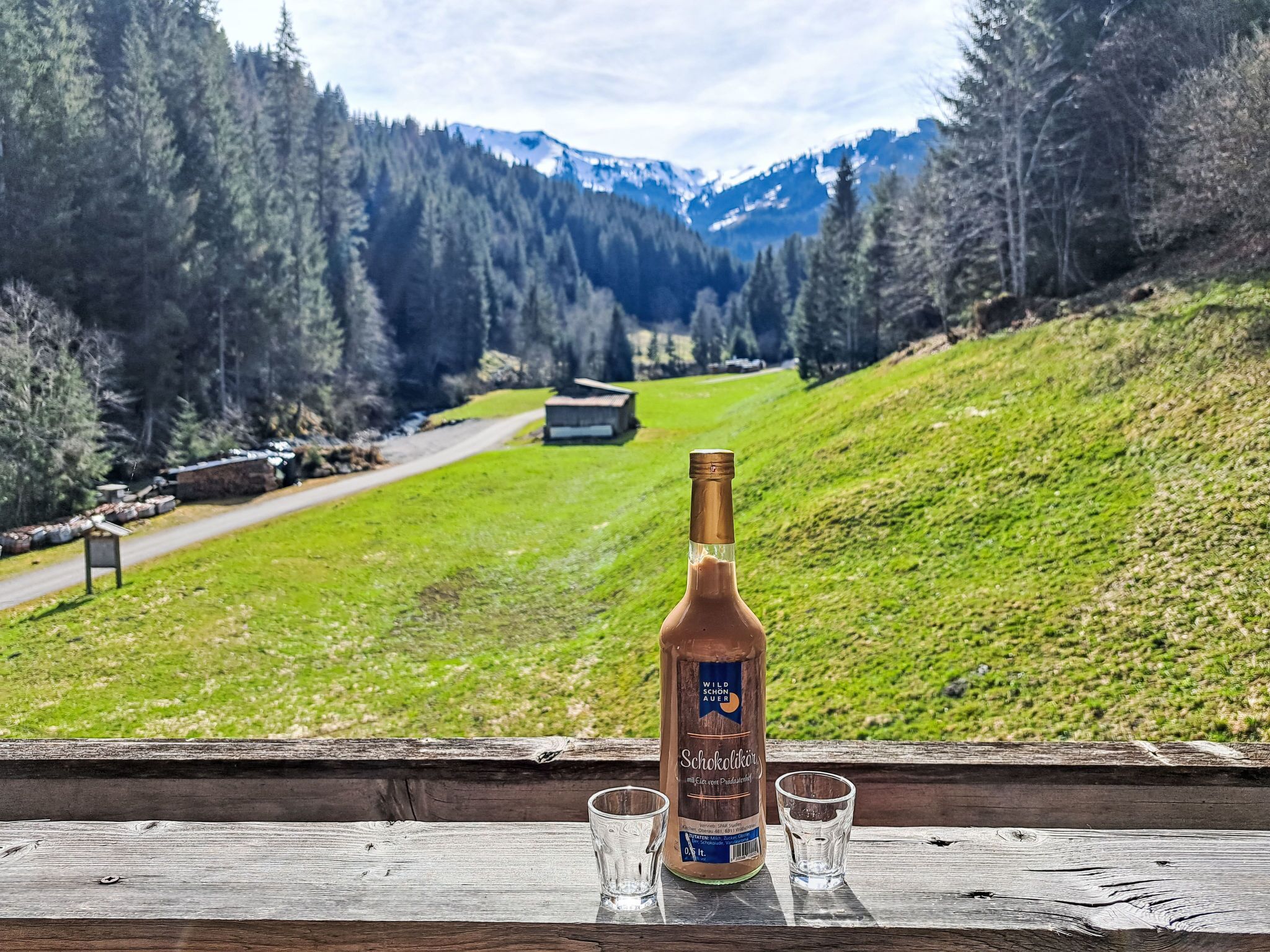 Una botella de Wild Schönauer Schokoladlikör y dos vasos sobre un alféizar de madera con vistas a un paisaje montañoso.