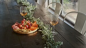 A long wooden table with a charcuterie board and two glasses of white wine, surrounded by white chairs and a window with a view of greenery. - Texas Sunshine Oasis WACO/Silos/Baylor: Pool, Piano, PingPong, Pets on 1/2 acre (Hewitt)