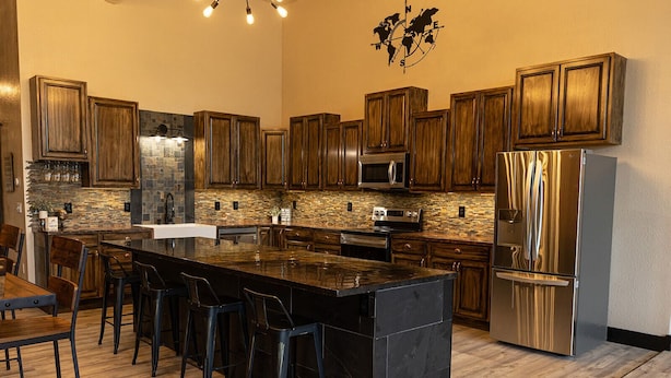 A modern kitchen with dark wooden cabinets, a large island with bar stools, a stainless steel refrigerator, and a stone backsplash.