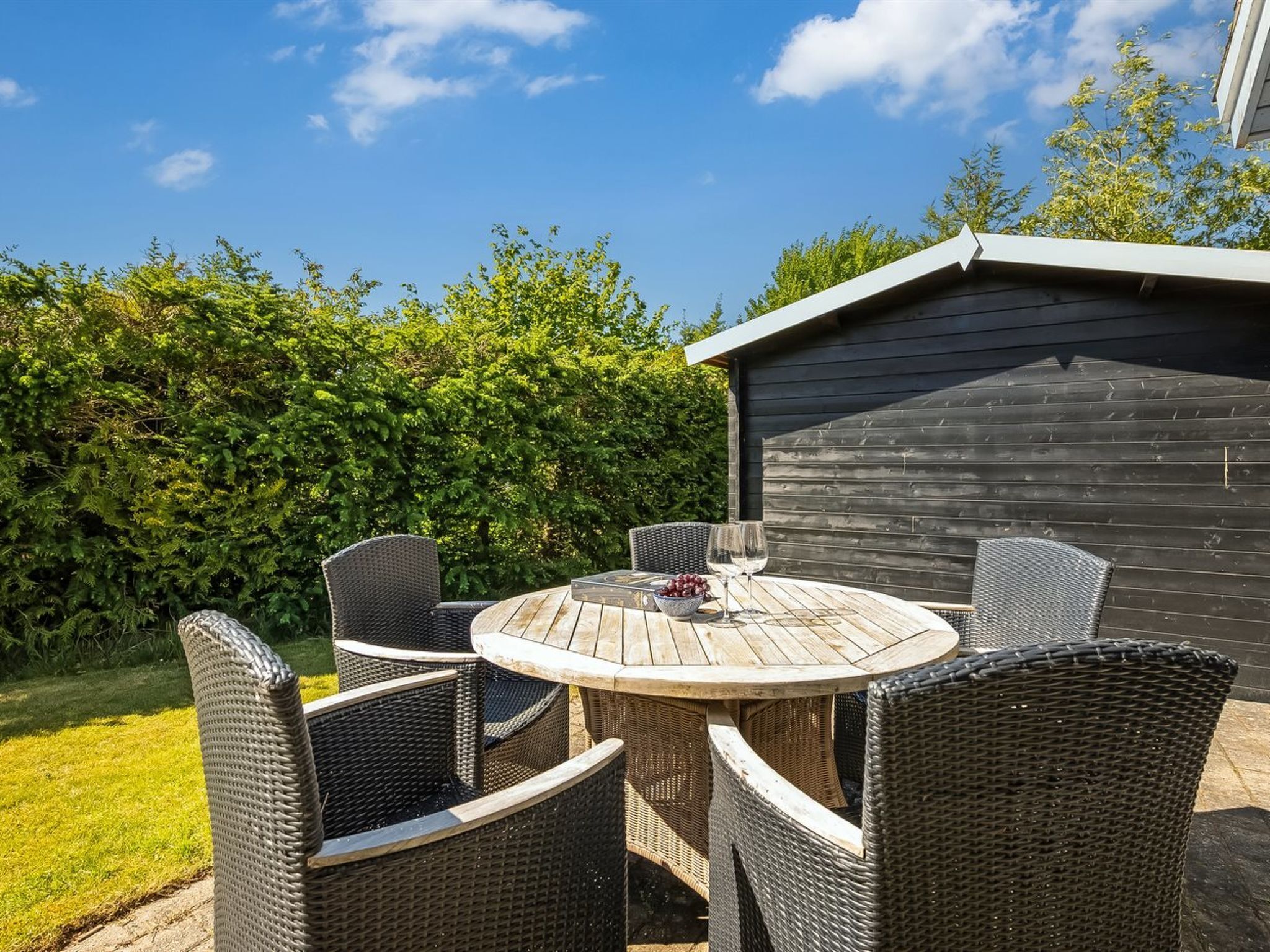 Eine Terrasse im Freien mit einem runden Tisch und Korbstühlen, umgeben von Grünpflanzen und einem Gebäude.