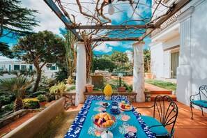 Une table dressée pour le petit-déjeuner dans un espace extérieur abrité, avec vue sur un bâtiment blanc et une végétation luxuriante.