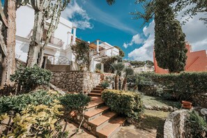 Une cour avec un escalier en pierre menant à un bâtiment blanc doté de balcons, entouré d’une végétation luxuriante et surplombé d’un ciel bleu clair.