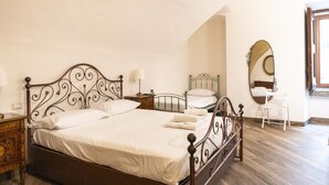 Een net opgemaakt bed met een metalen hoofdbord, een nachtkastje met een lamp, een kleine stoel en een spiegel aan de muur.