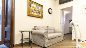 Sala de estar com sofá bege, mesa redonda pequena, quadro na parede e um relógio.