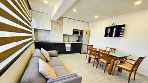 A modern living room with a grey sofa, wooden chairs, a small dining table, and a kitchen area with a microwave and a TV. - Centro Expositor, Feria, Estadios, Acuario, Planetario (Heroica Puebla de Zaragoza)