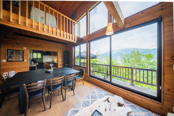 A wooden cabin with a dining area, kitchen, and large windows offering a mountain view.