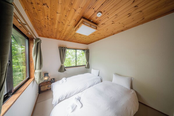 A room with two beds, a window with curtains, a bedside table with a lamp, and a wooden ceiling. - Wakuraba (Hakuba)