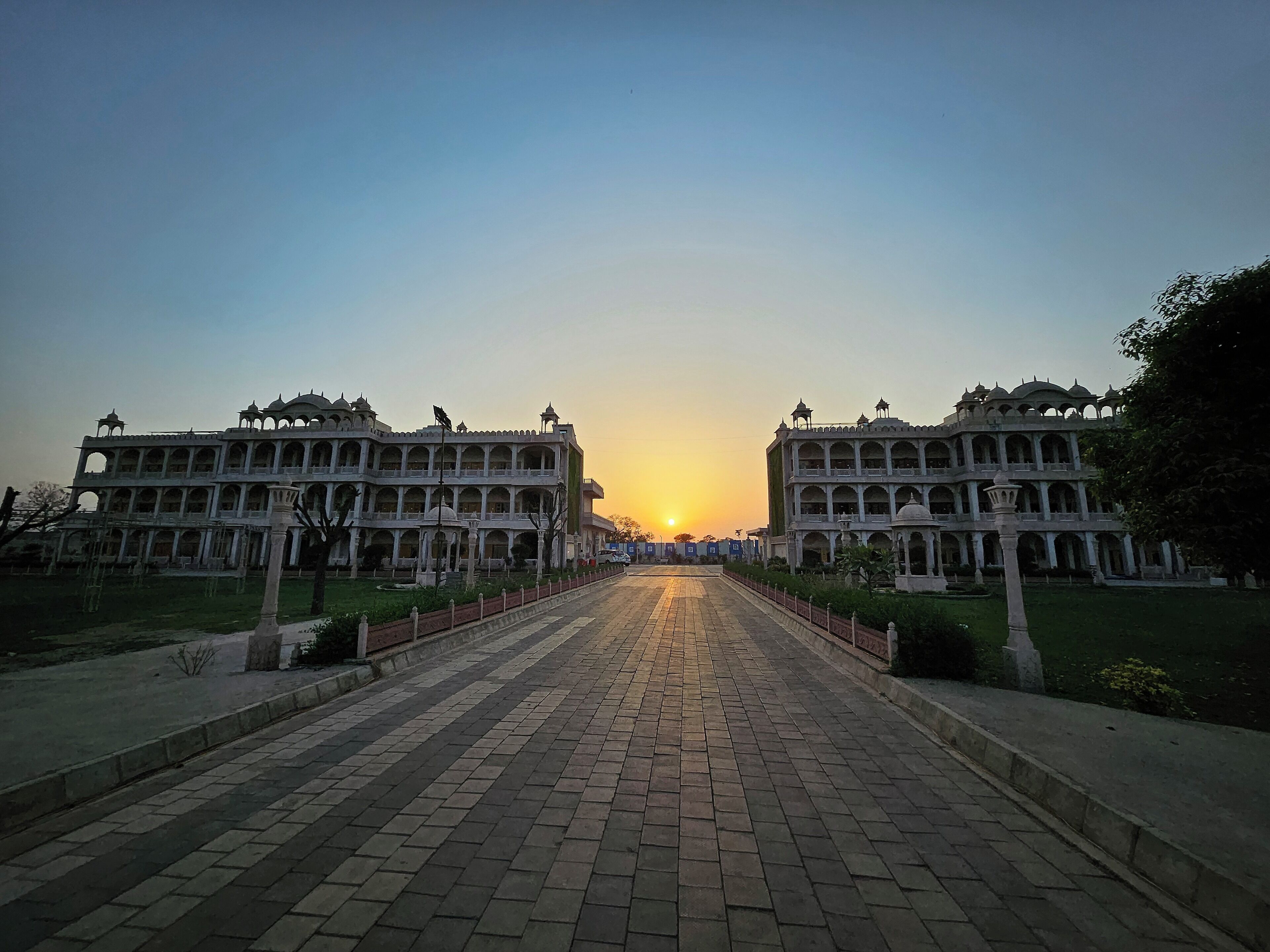 A grand building with multiple arches and domes, a paved pathway leading to it, and a sunset in the background.
