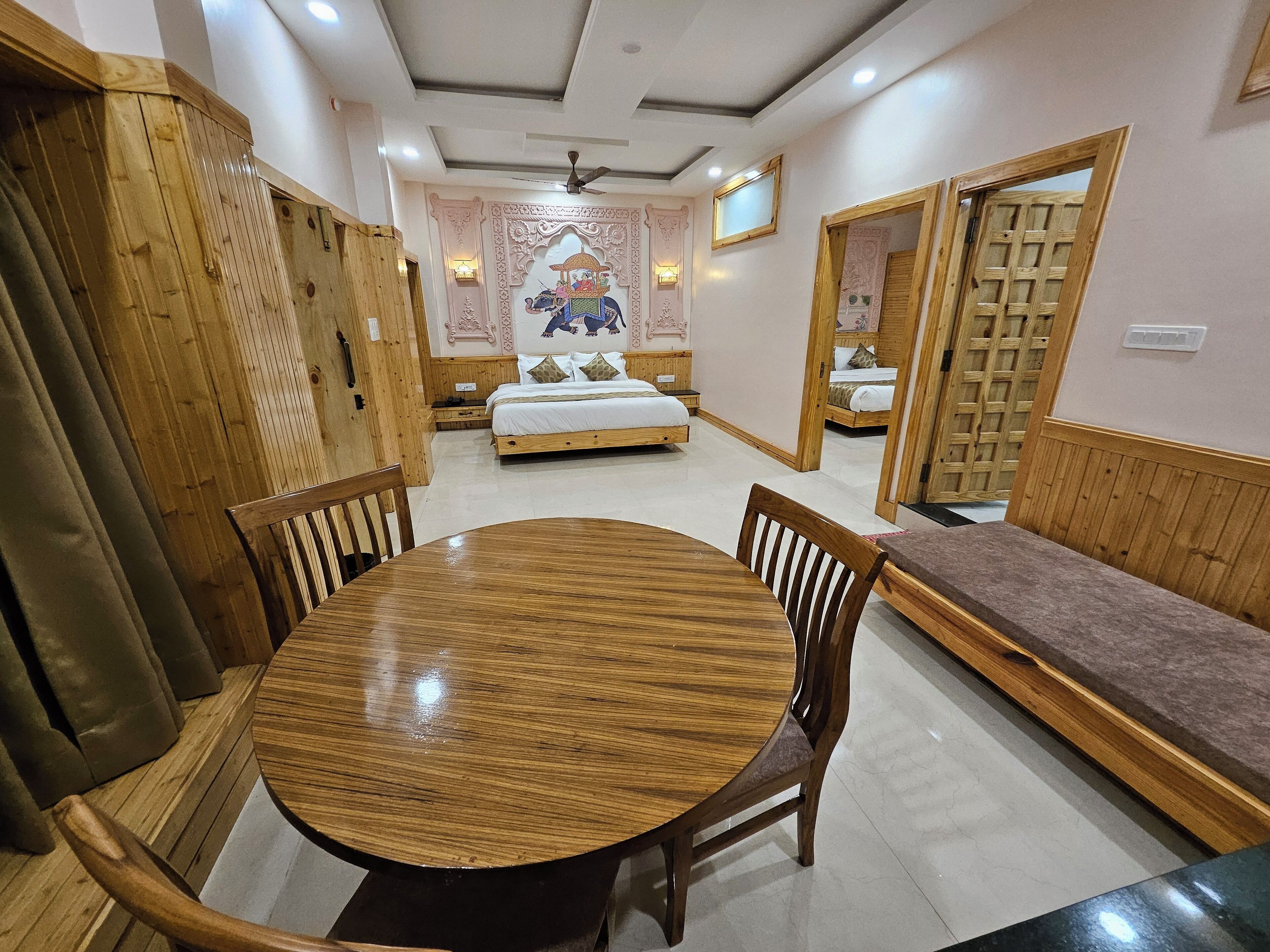 A room with a round wooden table, two wooden chairs, a bench, and a bed with a decorative wall mural.