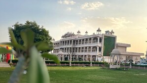 A large, historic building with multiple arches and domes, surrounded by a well-maintained lawn and a few parked cars. - The Mewar Palace and Resort (Shivpuri)