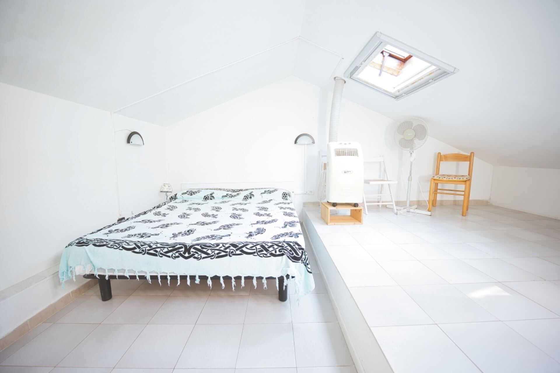 Un dormitorio con cama, una silla, un escritorio, un ventilador de pie y un tragaluz.