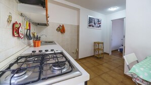 Eine Küche mit einem Gasherd, einem Spülbecken und verschiedenen Küchenutensilien. Im Raum befinden sich ein kleiner Holzschemel und ein Stuhl.