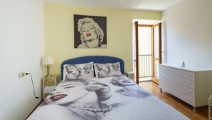 Ein Schlafzimmer mit einem Bett, das ein großes Marilyn-Monroe-Bild zeigt, einer Nachttischlampe, einem Schminktisch und einem Fenster mit Blick ins Freie.