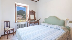 A bedroom with a bed, a chair, a dresser, and a window with a view of a town and a mountain. - Casa al secco apartment with sea view in the heart of Rio Marina (Rio Marina)