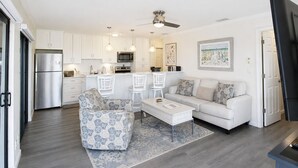 Een moderne keuken met witte kasten, een roestvrijstalen koelkast en een magnetron. Er is een eethoek met een witte tafel en stoelen, en een woonkamer met een bank en een gestreept fauteuil.