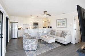 A modern kitchen with white cabinets, a stainless steel refrigerator, and a microwave. There is a dining area with a white table and chairs, and a living room with a sofa and patterned armchair.