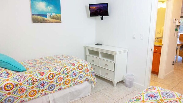 Un dormitorio con cama, televisión, cómoda blanca pequeña y baño visible a través de una puerta abierta.
