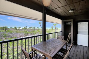 Une terrasse en bois avec une table et des chaises, un réfrigérateur blanc et une vue sur la plage et la végétation.