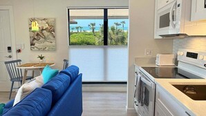 Cocina moderna con electrodomésticos blancos, zona de comedor con sofá azul y un amplio ventanal con vista a la playa.