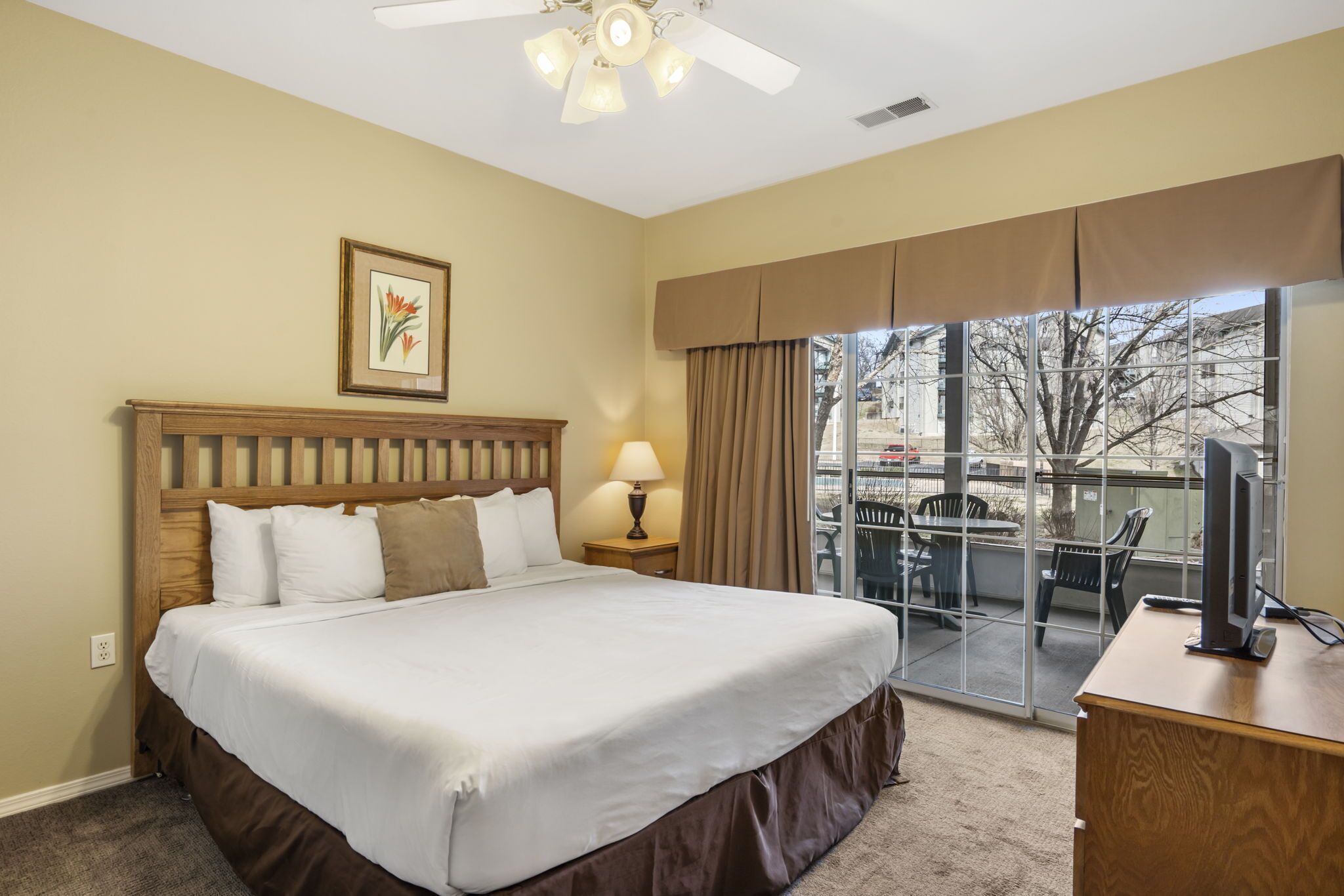 A hotel room with a large bed, a wooden headboard, a framed picture on the wall, a desk with a computer, and a balcony with outdoor furniture.