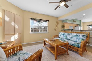 A living room with a sofa, coffee table, and chairs, a kitchen in the background, and a ceiling fan.