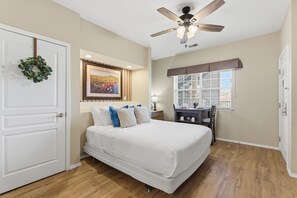 A bedroom with a bed, a ceiling fan, a desk, a window with blinds, and a door with a wreath.