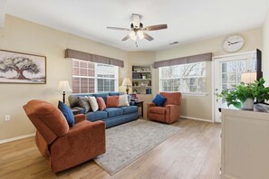 A living room with a blue sofa, two brown armchairs, a ceiling fan, a bookshelf, and a clock on the wall.