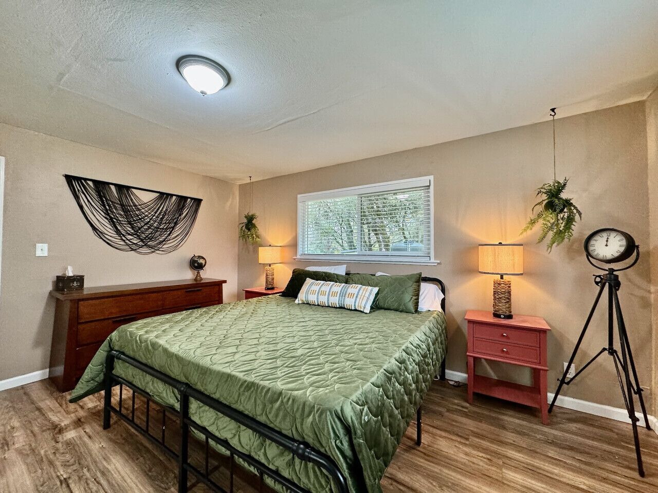 A bedroom with a green bedspread, wooden dresser, and a round clock on a tripod stand.