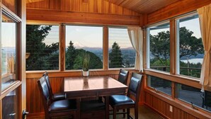 Un'area pranzo con tavolo e sedie in legno, ampie finestre vista su montagna e alberi.