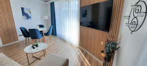 Una sala de estar moderna con un televisor de pantalla plana, una mesa redonda para café, dos sillas azules, un sofá y una lámpara.