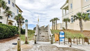 Une entrée de plage avec un escalier en bois menant à une zone de dunes, des palmiers et un panneau indiquant : « Reconstruction des dunes, accès interdit ».
