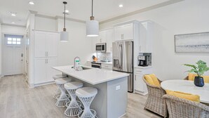 Une cuisine moderne avec des meubles blancs, un îlot central doté de tabourets de bar, des appareils en acier inoxydable et un coin repas avec des meubles en osier.
