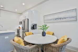 A modern kitchen with white cabinetry, stainless steel appliances, and a dining area with wicker chairs and a round table. - Beautiful condo with pool, hot tub, beach access, central AC, & washer/dryer (Santa Rosa Beach)