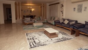 A spacious living room with a large sofa, ottoman, and a coffee table. There are two chairs, a chandelier, and framed artwork on the walls. - villa in deplomaseen district (Sheikh Zayed City)