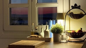 A kitchen with a wooden countertop, a window with a view of the sunset, and a basket of fruits.