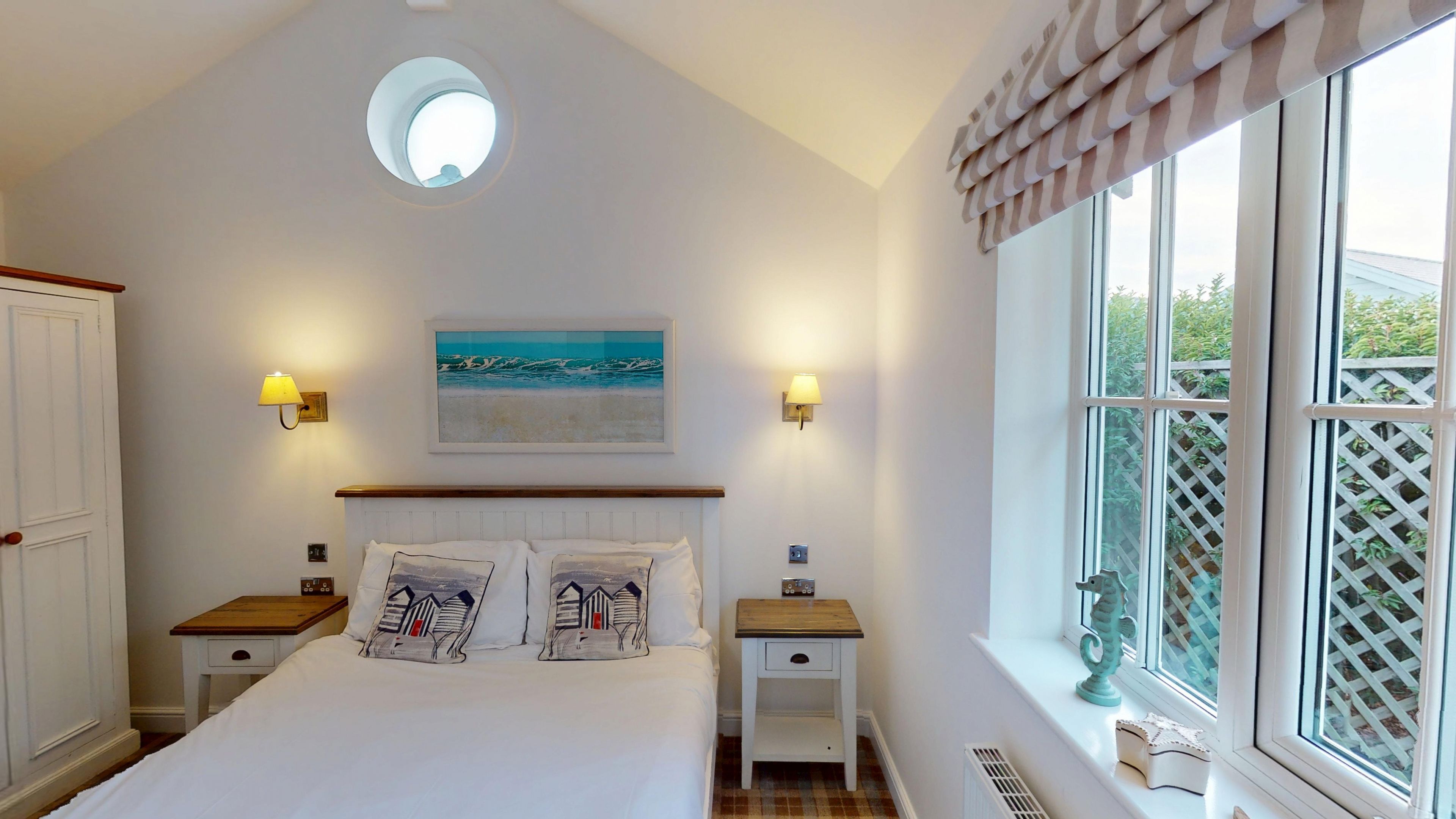 Een slaapkamer met een bed, twee nachtkastjes, een wandlamp, een plafondlamp, een raam met uitzicht op groen en een ingelijst schilderij aan de muur.